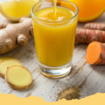 belebender ingwer zitronen shot mit kurkuma - Ein goldener, belebender Ingwer-Zitronen-Shot mit Kurkuma in einem kleinen Glas, garniert mit Zitronenscheiben und Ingwerstücken, im Hintergrund frische Ingwer-, Kurkuma- und Zitronenscheiben auf einem Holzbrett.