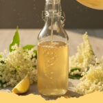 frischer holunderbluetensirup selbst gemacht - Eine Reihe von Glasflaschen, gefüllt mit goldgelbem, frischem Holunderblütensirup, umgeben von frischen Holunderblütendolden und Zitronenscheiben auf einem rustikalen Holztisch.