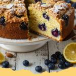 luftiger zitronen blaubeer kuchen mit mandelstreusel - Ein großes Stück luftiger Zitronen-Blaubeer-Kuchen mit Mandelstreuseln auf einem Teller, daneben frische Blaubeeren und Zitronenscheiben.